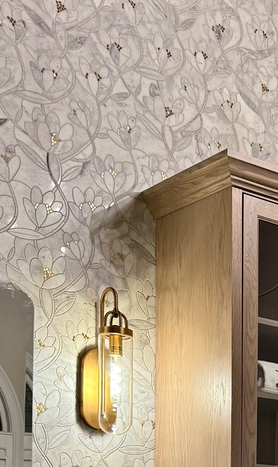 Marble mosaic floral accent wall with a brass wall sconce beside a natural wood cabinet